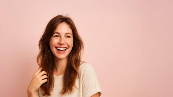 Jonge vrouw met gezond, glanzend haar die lachend haar hand door haar haar haalt tegen een pastelroze achtergrond.
