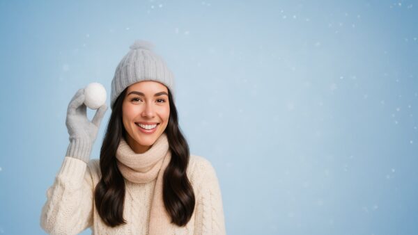 Blije jonge vrouw met glanzend haar in trui, sjaal en muts houdt speels een sneeuwbal vast tegen een pastel-winternacht-achtergrond.