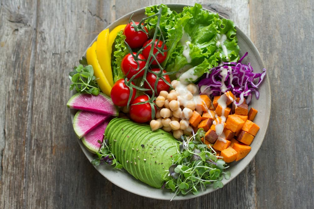 Een kleurrijke salade bowl met avocado, kikkererwten, zoete aardappel, kerstomaatjes, sla, paprika, rode kool en watermeloenradijs, geserveerd met een lichte dressing.