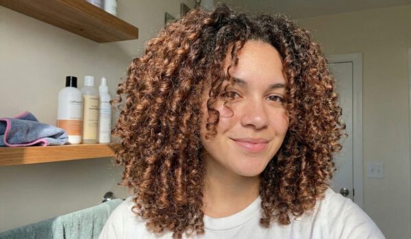 Vrouw met gezonde, gedefinieerde en pluisvrije krullen in een badkamer met haarproducten op de achtergrond, als resultaat van de simpele krullen routine voor beginners.