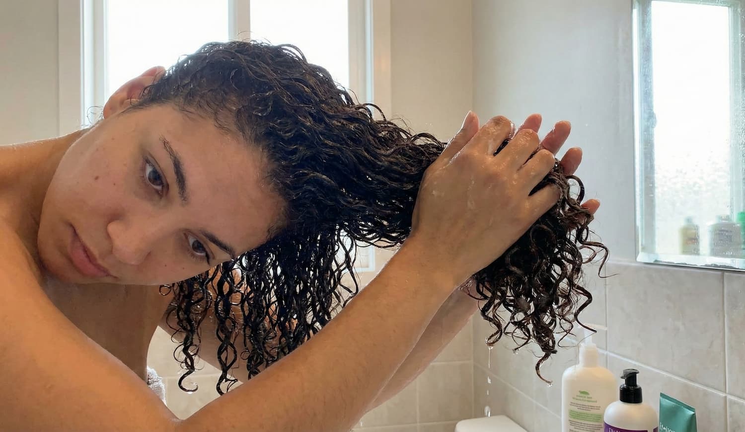 Een close-up van een vrouw met nat, donker krullend haar die haar hoofd schuin houdt in een badkamer. Ze demonstreert de 'scrunch' techniek door haar gedefinieerde krullen in haar handen op te vangen en ze omhoog richting haar hoofdhuid te knijpen. Op de achtergrond zijn een raam en flessen haarverzorgingsproducten te zien.