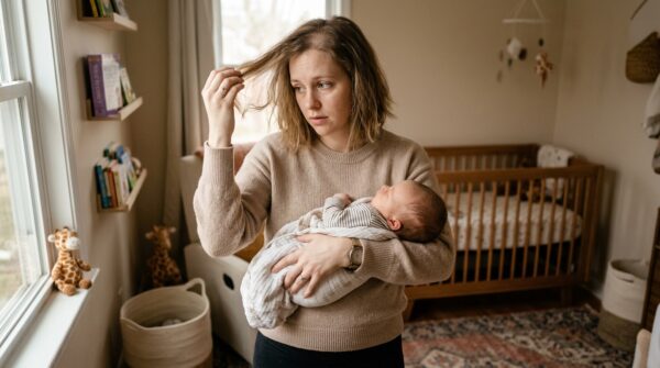 Een jonge moeder die een baby vasthoudt in een babykamer en met een bezorgde blik naar haar dunner wordende haar kijkt.
